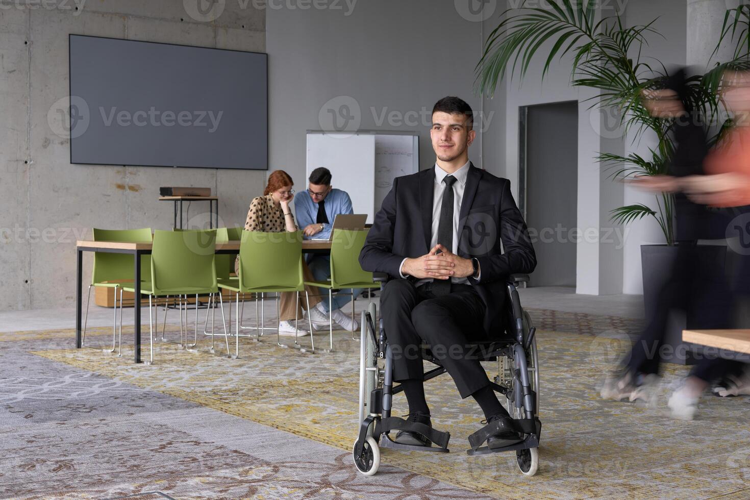 A businessman in a wheelchair navigating through a busy office, surrounded by his colleagues who are actively engaged in their work and collaboration photo