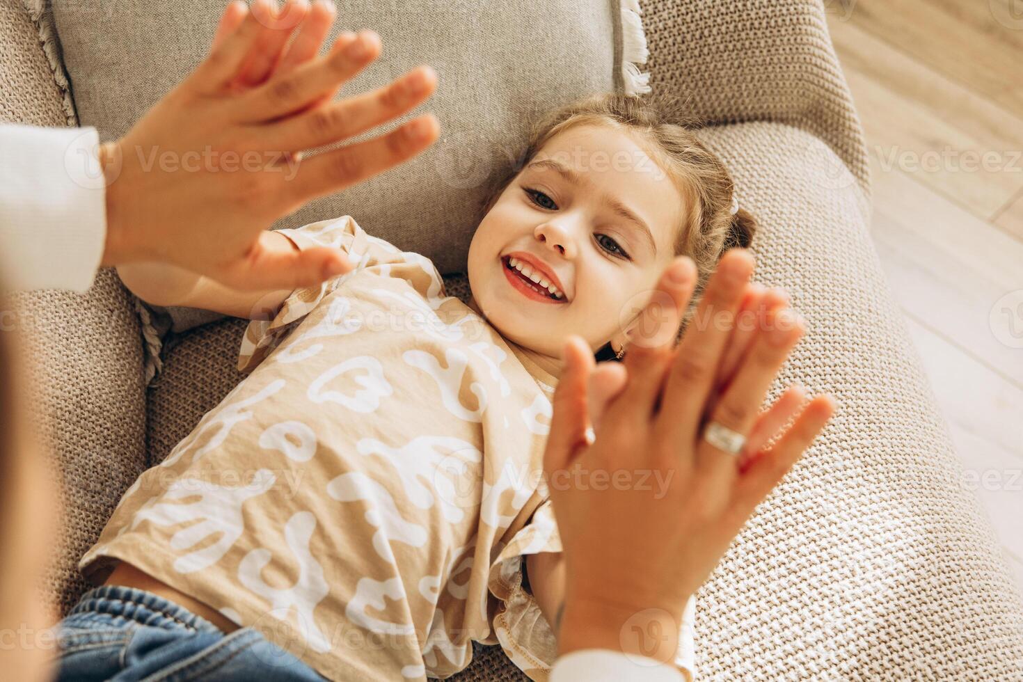 Smiling little daughter clapping hands with mother lying on comfortable sofa in living room, at home photo