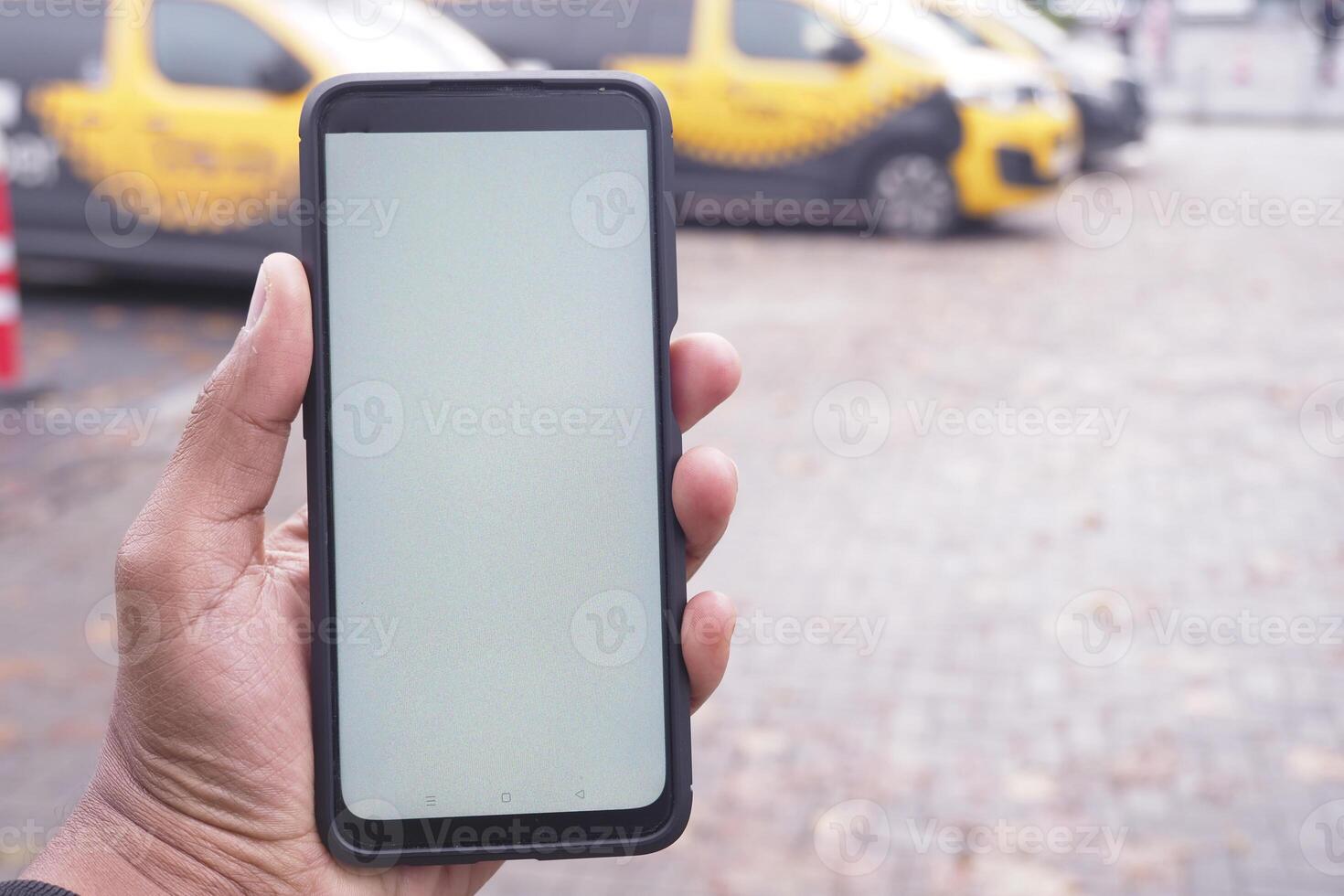A hand is holding a smartphone that features a blank screen amidst yellow taxis in the background photo