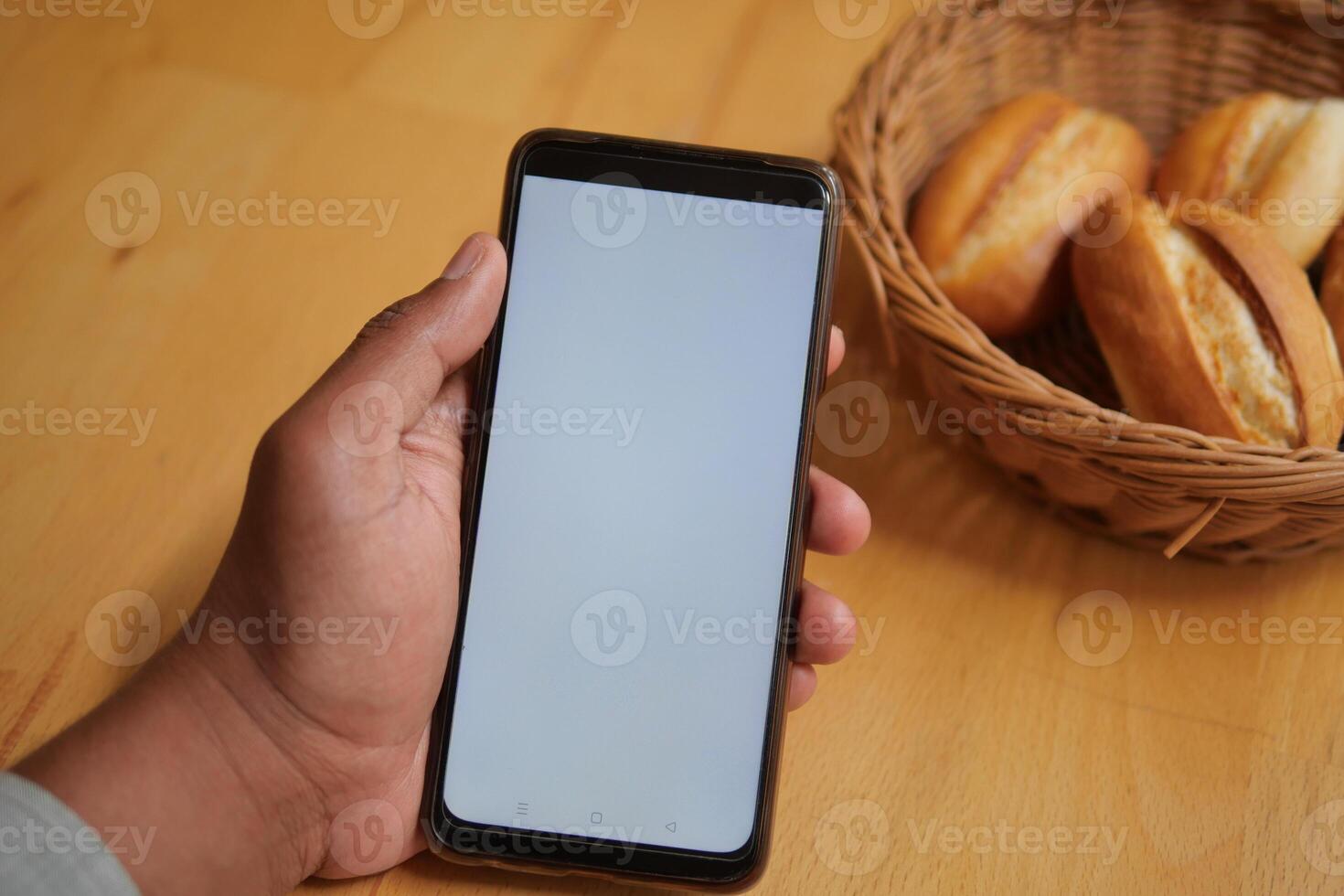 A hand is holding a smartphone that has an empty screen beside some freshly baked buns photo