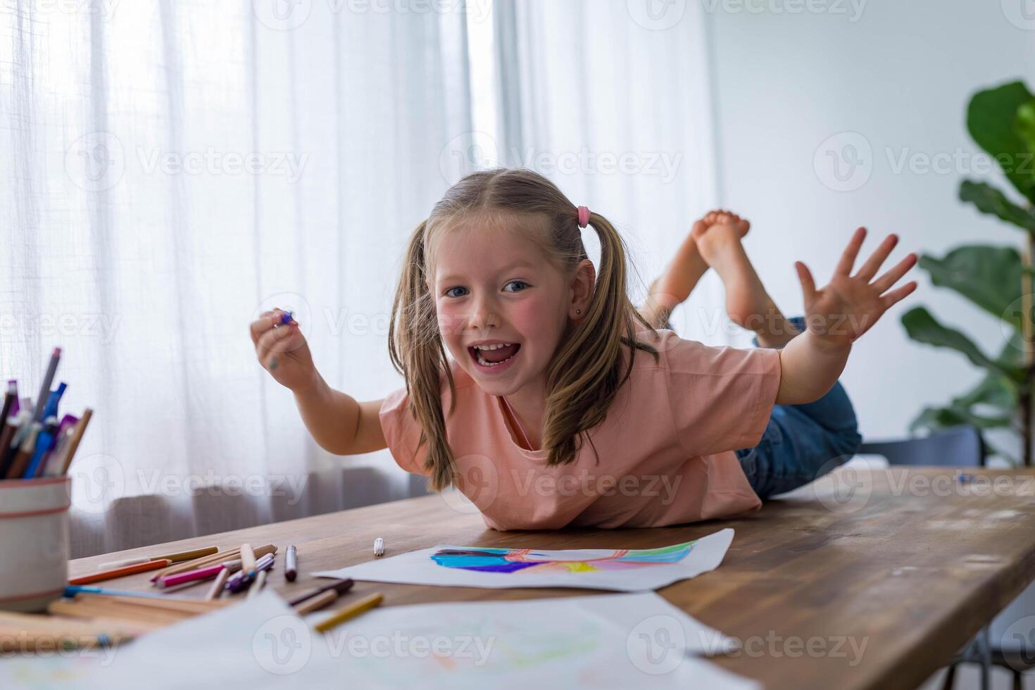 A 5-year-old girl laughs joyfully as she draws at the table, surrounded by colorful pencils photo
