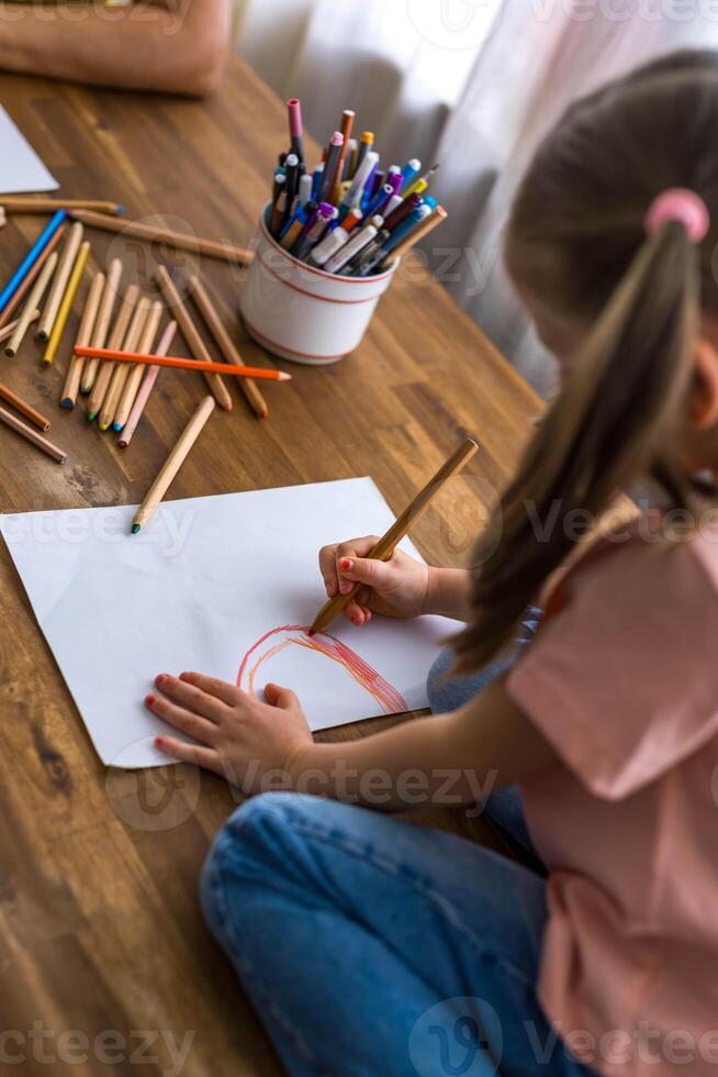 A 5-year-old girl happily draws at a table surrounded by a rainbow of colorful pencils,top view photo