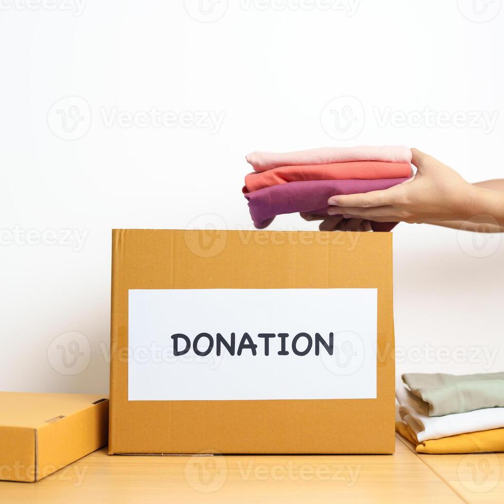 Donation, Charity, Volunteer, Giving and Delivery Concept. Hand holding Clothes into Donation box at home or office for support and help poor, refugee and homeless people. Copy space for text photo
