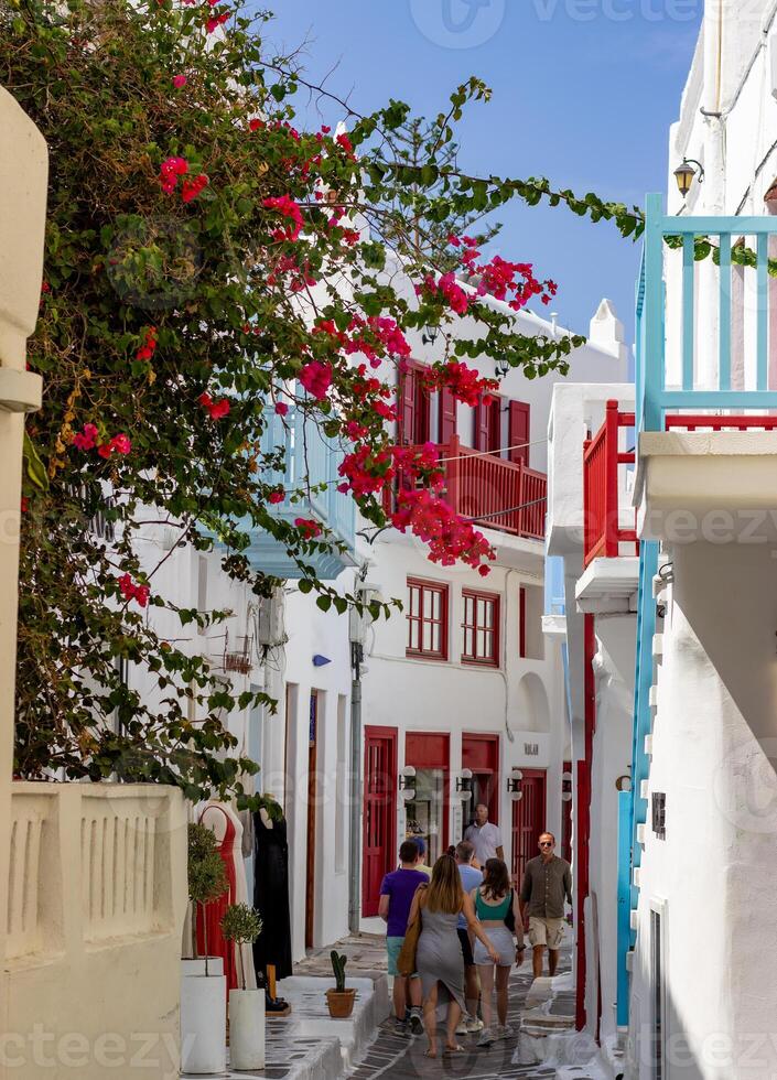 Narrow street in Mykonos with whitewashed buildings, colorful balconies, and blooming bougainvillea. People walking in a traditional Greek village. photo