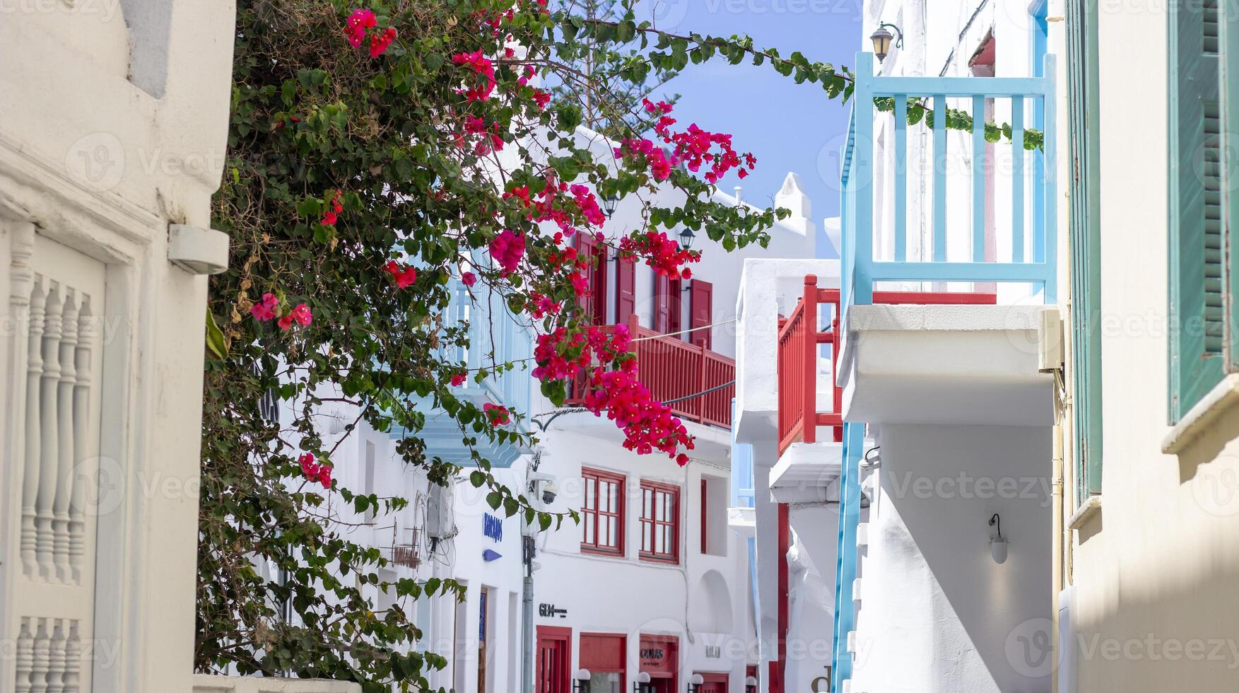 Narrow street in Mykonos with whitewashed buildings, colorful balconies, and blooming bougainvillea. People walking in a traditional Greek village. photo