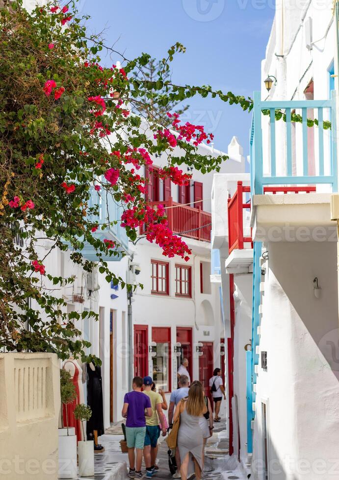 Narrow street in Mykonos with whitewashed buildings, colorful balconies, and blooming bougainvillea. People walking in a traditional Greek village. photo