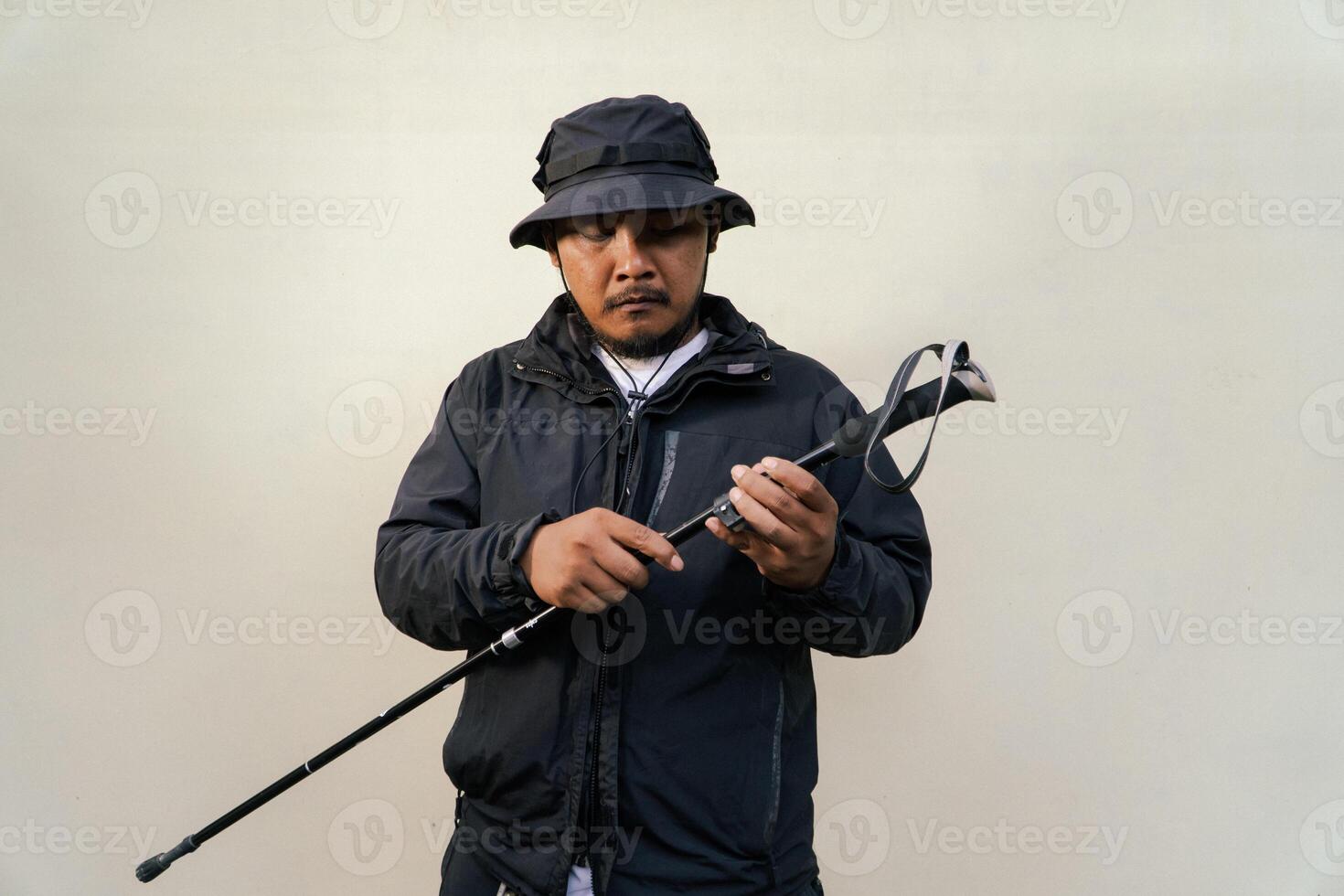 Photo of Asian man wearing adventuring outfit. Half body portrait of an adventurer with beard and mustache posing climber wearing jacket, boonie hat and trekking poles isolated on beige background