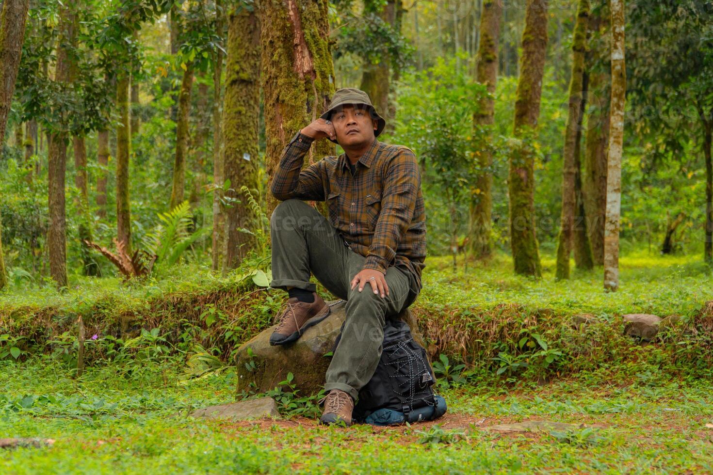 An adventurer is sitting on a large rock in a dense pine forest. An Asian man in a flannel shirt and bucket hat is sitting and resting while enjoying the tranquility of the beautiful nature. photo