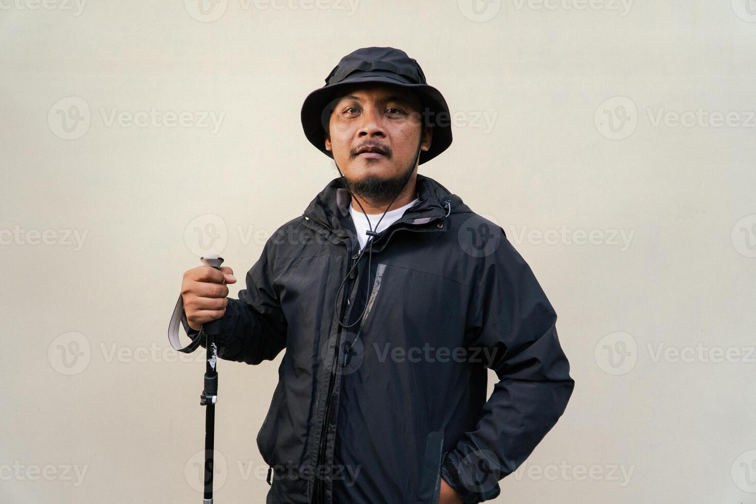 Photo of Asian man wearing adventuring outfit. Half body portrait of an adventurer with beard and mustache posing climber wearing jacket, boonie hat and trekking poles isolated on beige background