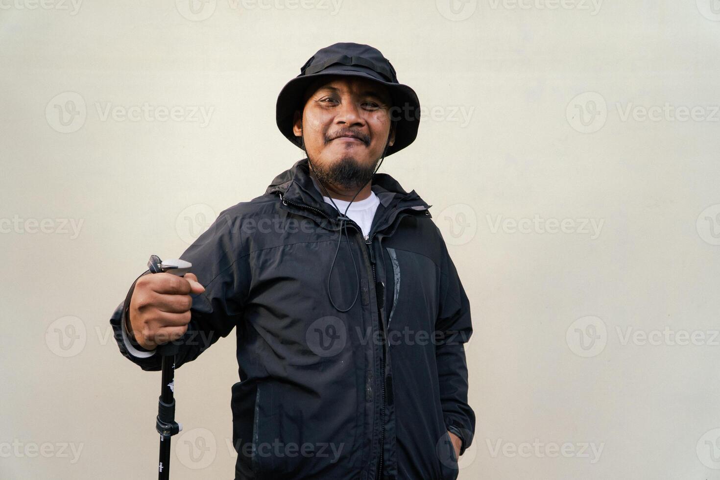 Photo of Asian man wearing adventuring outfit. Half body portrait of an adventurer with beard and mustache posing climber wearing jacket, boonie hat and trekking poles isolated on beige background