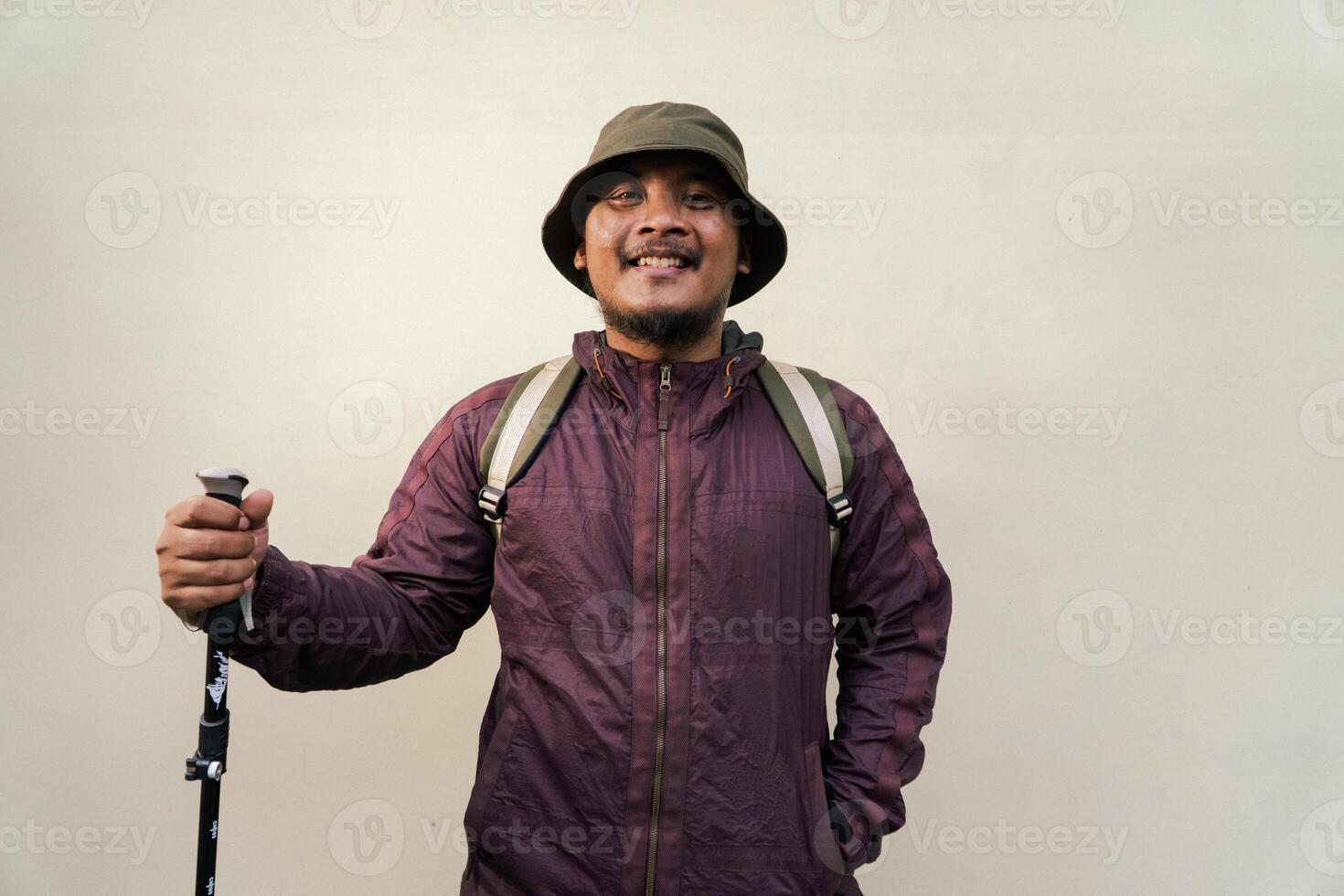 Adult Asian man wearing adventurer outfit and posing in various poses. An adventurer wearing maroon jacket, bucket hat, backpack and trekking pole isolated on beige background. photo