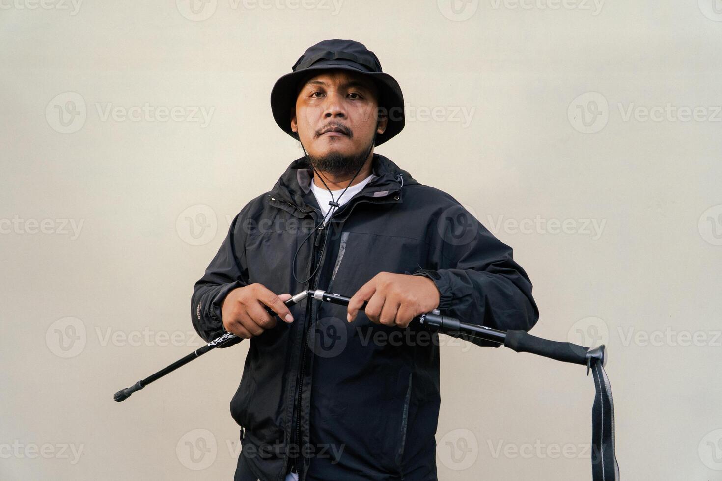 Photo of Asian man wearing adventuring outfit. Half body portrait of an adventurer with beard and mustache posing climber wearing jacket, boonie hat and trekking poles isolated on beige background