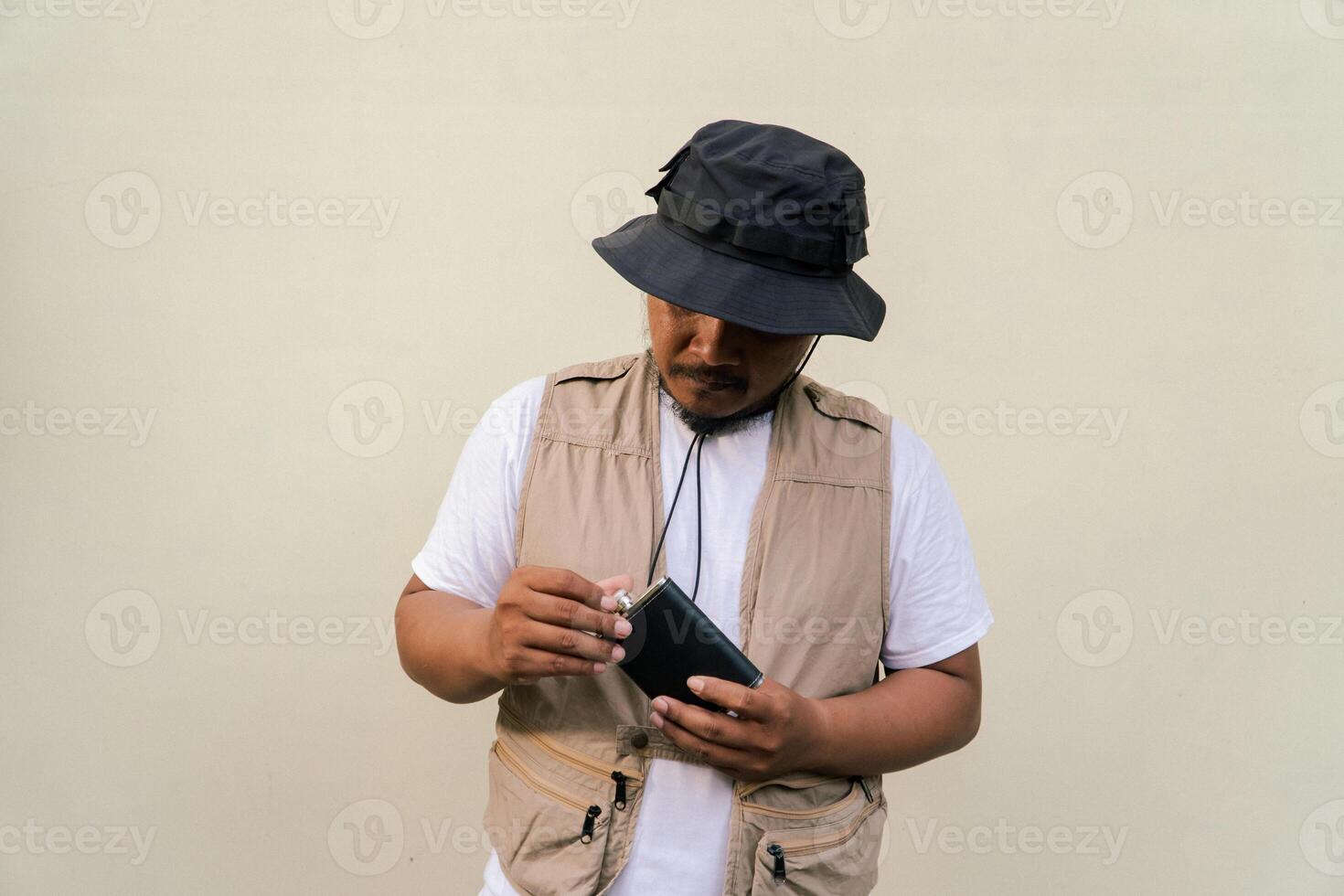 Macho mature man with beard and mustache wearing safari outfit isolated on beige background. Half body portrait of an adult Southeast Asian man posing wearing a vest, bucket hat and hip flask bottle photo