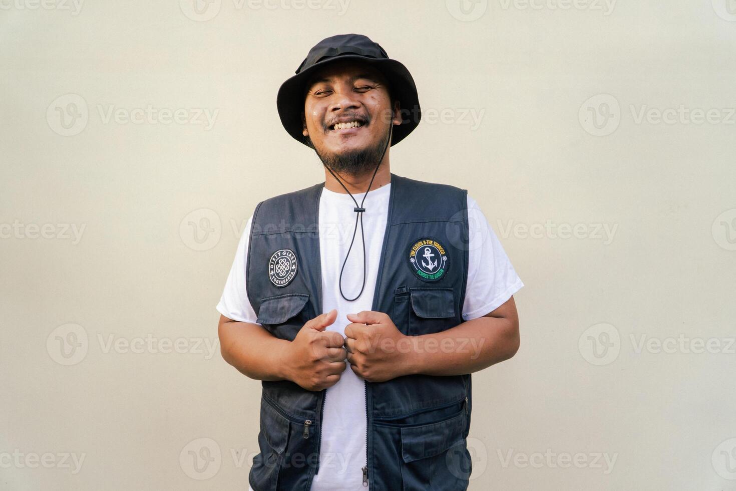 Asian adult man wearing safari suit isolated on beige background. Half body portrait of adult Southeast Asian man posing wearing vest, bucket hat and white t-shirt photo