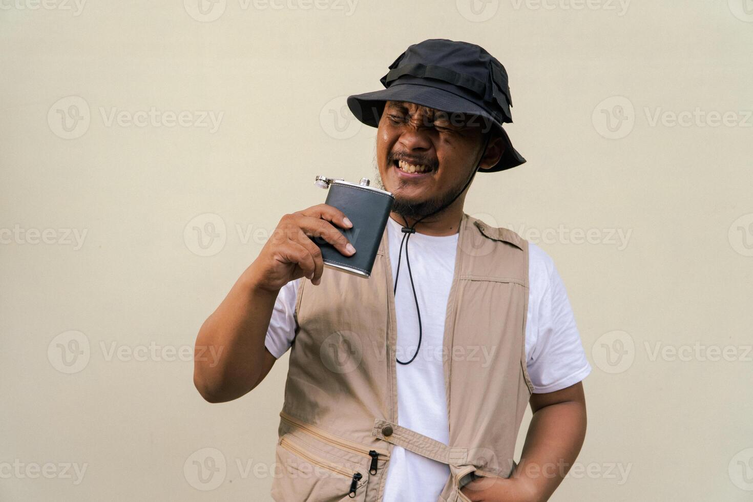 Macho mature man with beard and mustache wearing safari outfit isolated on beige background. Half body portrait of an adult Southeast Asian man posing wearing a vest, bucket hat and hip flask bottle photo