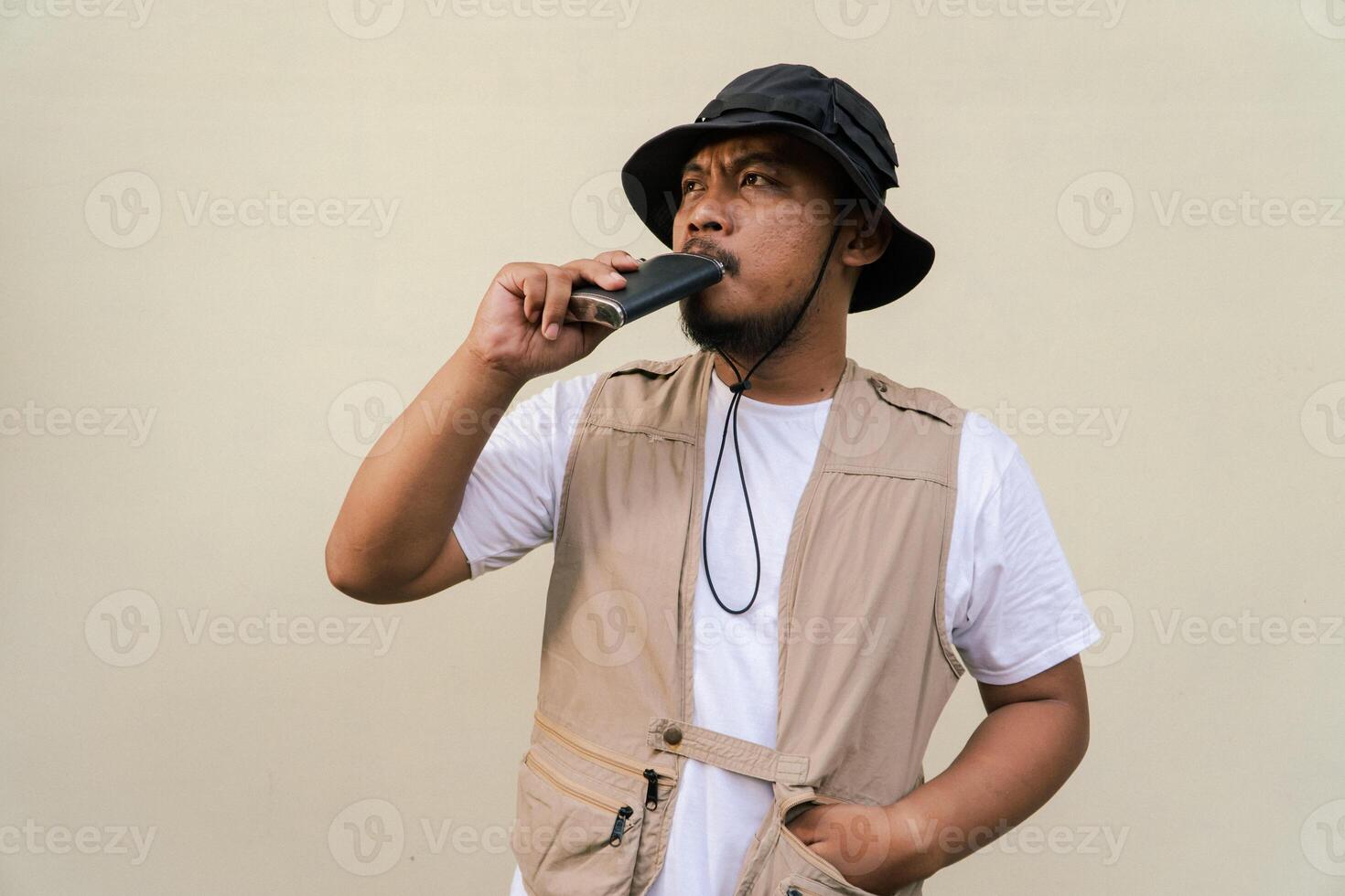Macho mature man with beard and mustache wearing safari outfit isolated on beige background. Half body portrait of an adult Southeast Asian man posing wearing a vest, bucket hat and hip flask bottle photo