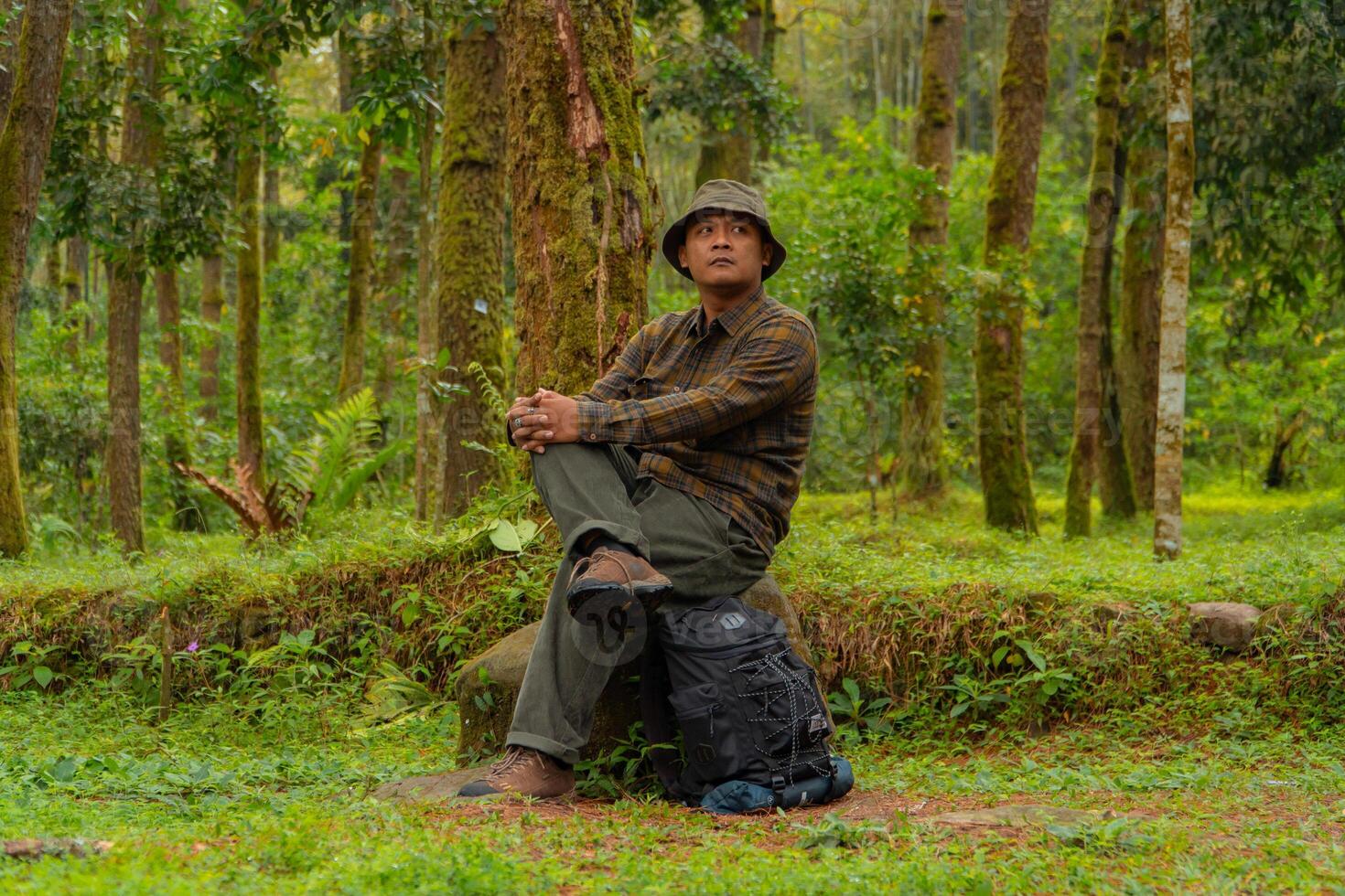 An adventurer is sitting on a large rock in a dense pine forest. An Asian man in a flannel shirt and bucket hat is sitting and resting while enjoying the tranquility of the beautiful nature. photo