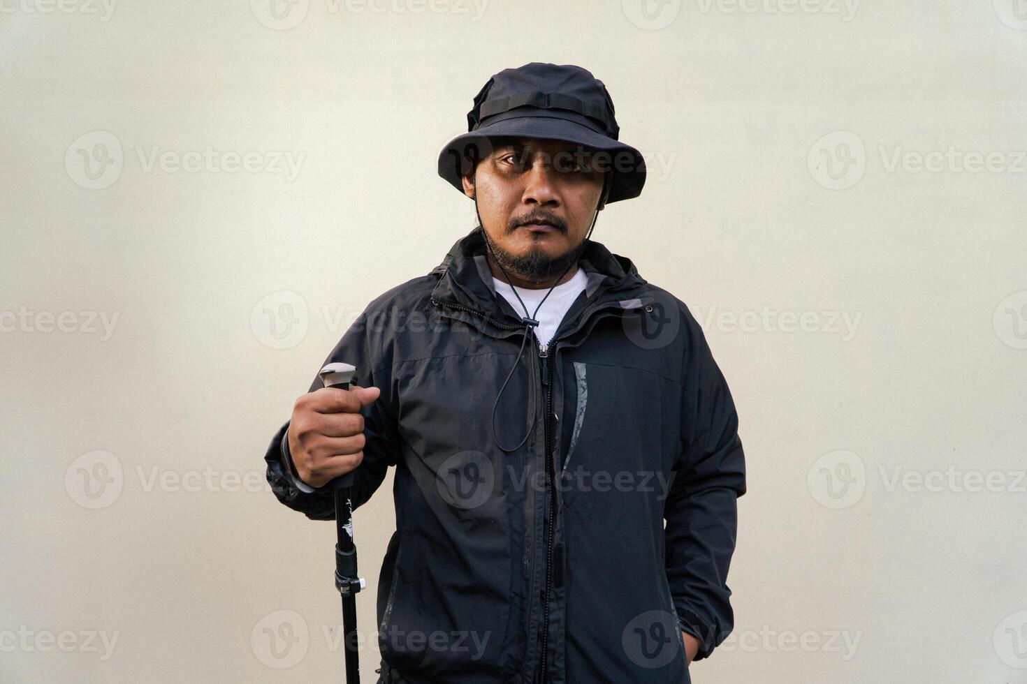 Photo of Asian man wearing adventuring outfit. Half body portrait of an adventurer with beard and mustache posing climber wearing jacket, boonie hat and trekking poles isolated on beige background