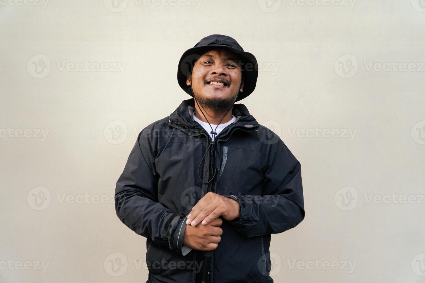 Photo of Asian man wearing adventuring outfit. Half body portrait of an adventurer with beard and mustache posing climber wearing jacket, boonie hat and trekking poles isolated on beige background