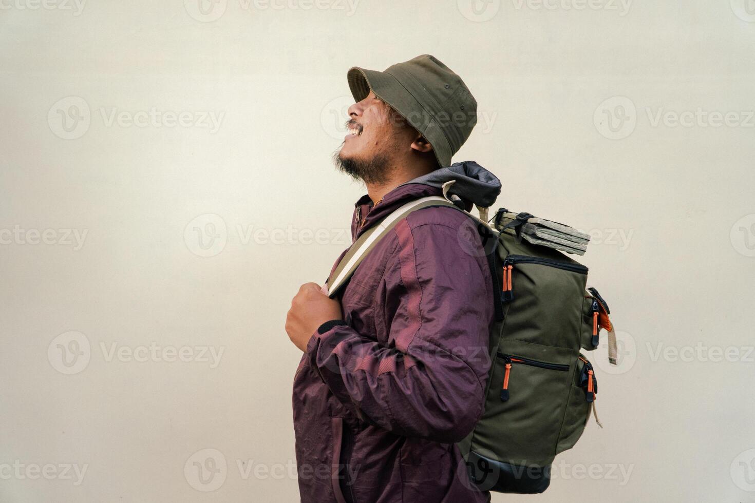 Half body portrait of adult Asian man with beard and mustache posing and wearing mountain climbing outfit with jacket, bucket hat, carrier bag and trekking poles isolated on beige background. photo