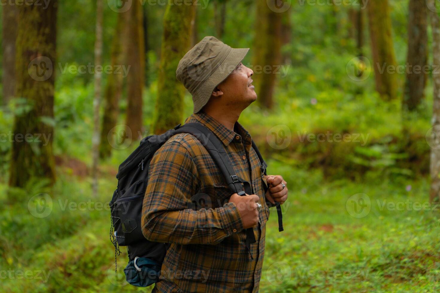 Half-body close-up of an adventurer in a dense and green pine forest. An Asian male traveler in a flannel shirt, bucket hat and backpack poses against a backdrop of dense pine trees. photo