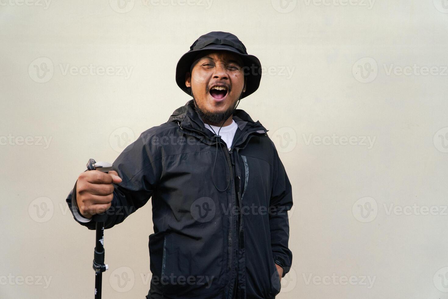 Photo of Asian man wearing adventuring outfit. Half body portrait of an adventurer with beard and mustache posing climber wearing jacket, boonie hat and trekking poles isolated on beige background