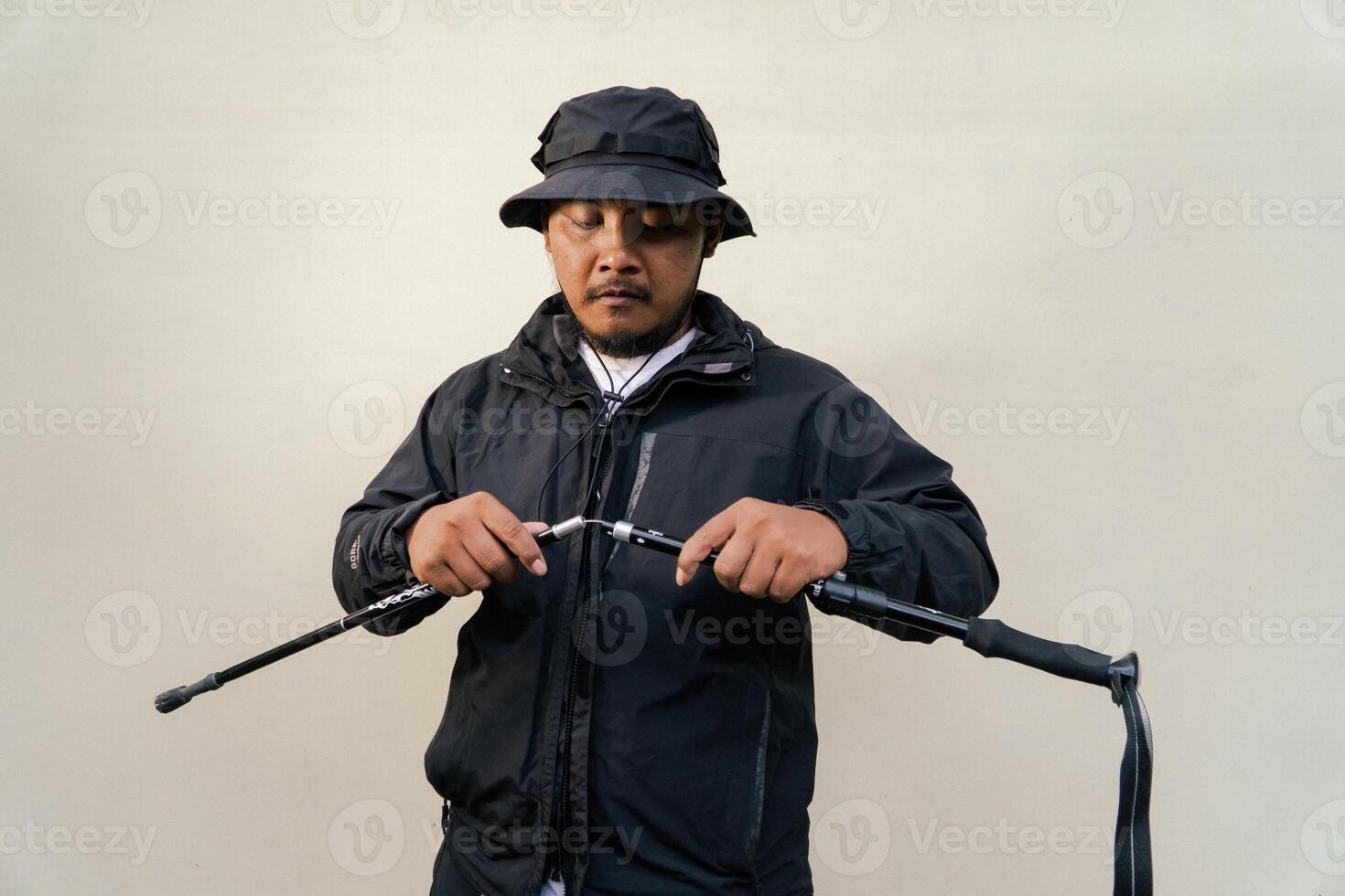 Photo of Asian man wearing adventuring outfit. Half body portrait of an adventurer with beard and mustache posing climber wearing jacket, boonie hat and trekking poles isolated on beige background