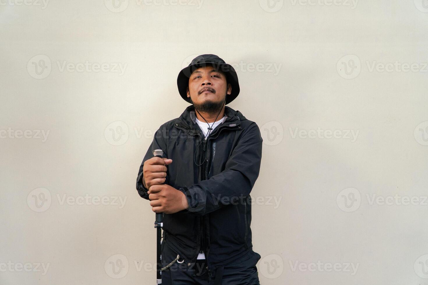 Photo of Asian man wearing adventuring outfit. Half body portrait of an adventurer with beard and mustache posing climber wearing jacket, boonie hat and trekking poles isolated on beige background