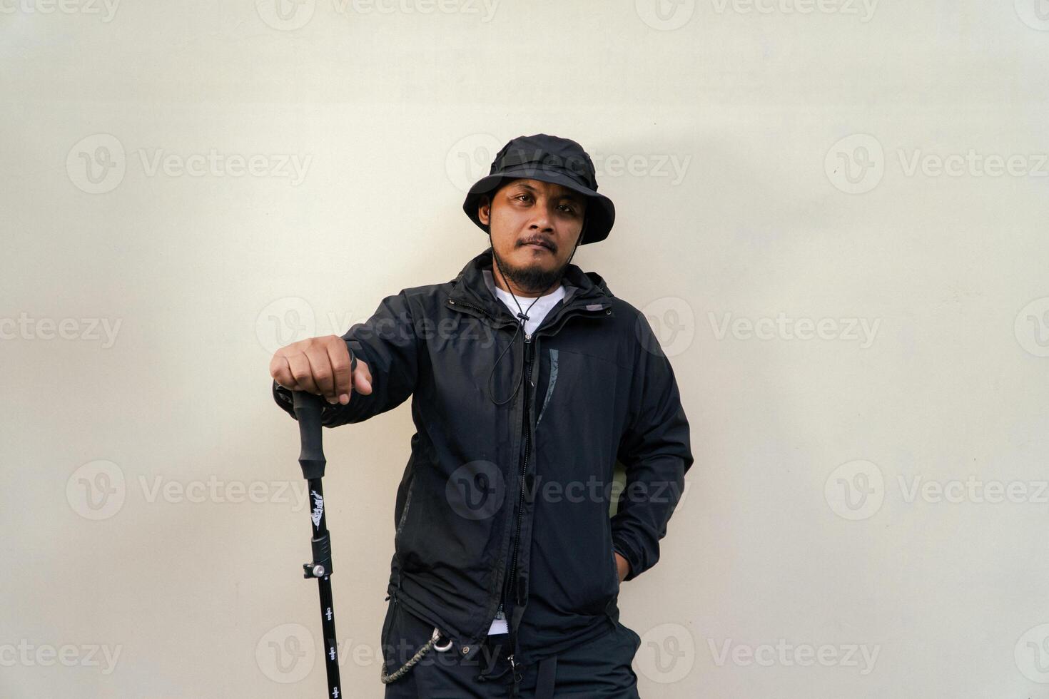 Photo of Asian man wearing adventuring outfit. Half body portrait of an adventurer with beard and mustache posing climber wearing jacket, boonie hat and trekking poles isolated on beige background