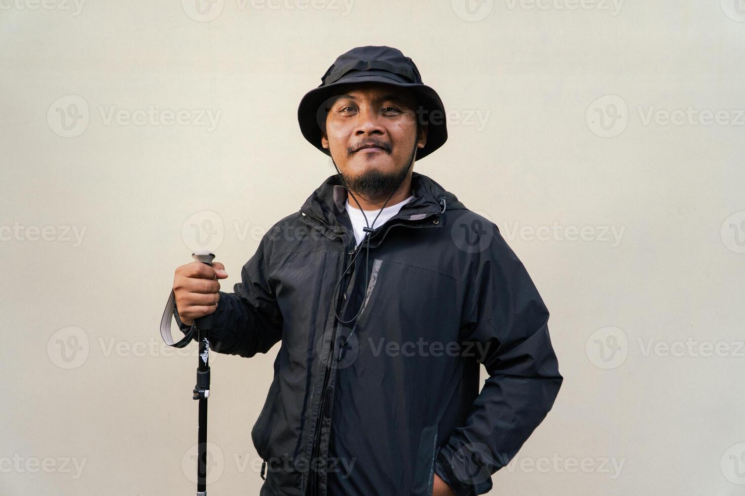 Photo of Asian man wearing adventuring outfit. Half body portrait of an adventurer with beard and mustache posing climber wearing jacket, boonie hat and trekking poles isolated on beige background