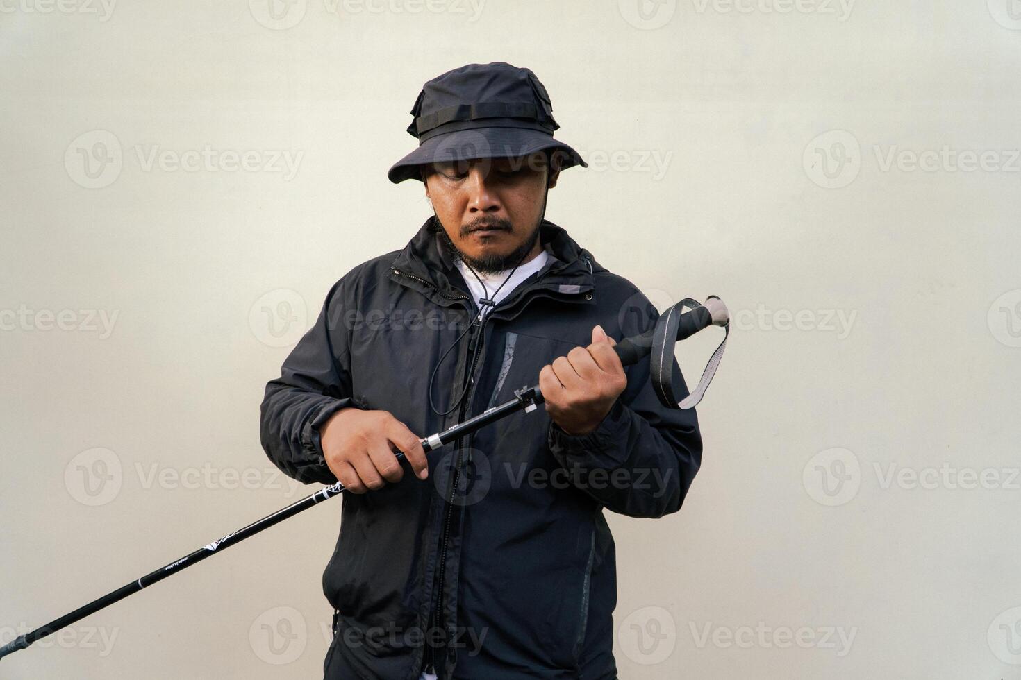 Photo of Asian man wearing adventuring outfit. Half body portrait of an adventurer with beard and mustache posing climber wearing jacket, boonie hat and trekking poles isolated on beige background