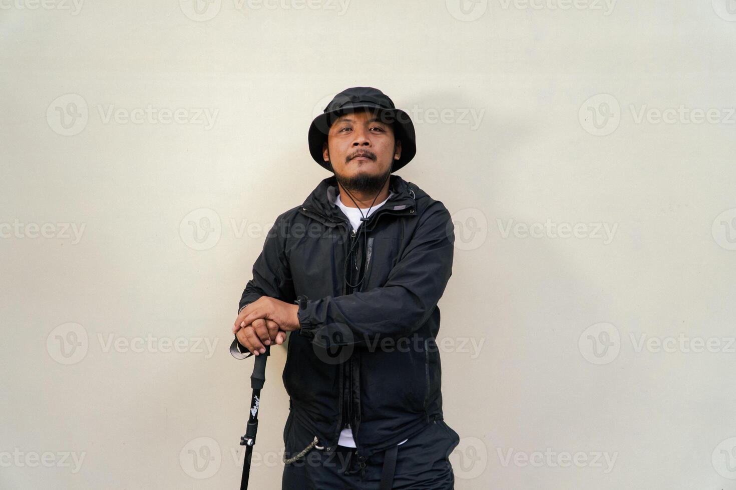 Photo of Asian man wearing adventuring outfit. Half body portrait of an adventurer with beard and mustache posing climber wearing jacket, boonie hat and trekking poles isolated on beige background