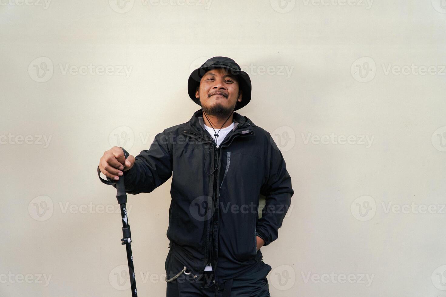 Photo of Asian man wearing adventuring outfit. Half body portrait of an adventurer with beard and mustache posing climber wearing jacket, boonie hat and trekking poles isolated on beige background