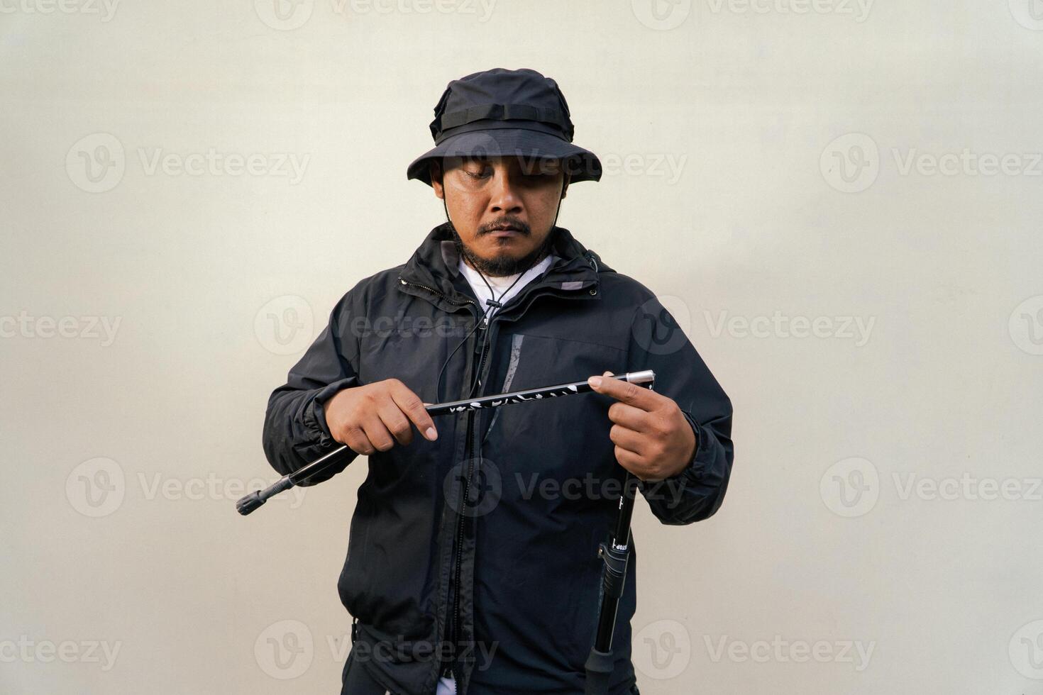 Photo of Asian man wearing adventuring outfit. Half body portrait of an adventurer with beard and mustache posing climber wearing jacket, boonie hat and trekking poles isolated on beige background