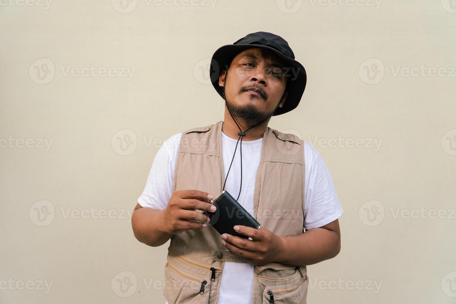 Macho mature man with beard and mustache wearing safari outfit isolated on beige background. Half body portrait of an adult Southeast Asian man posing wearing a vest, bucket hat and hip flask bottle photo