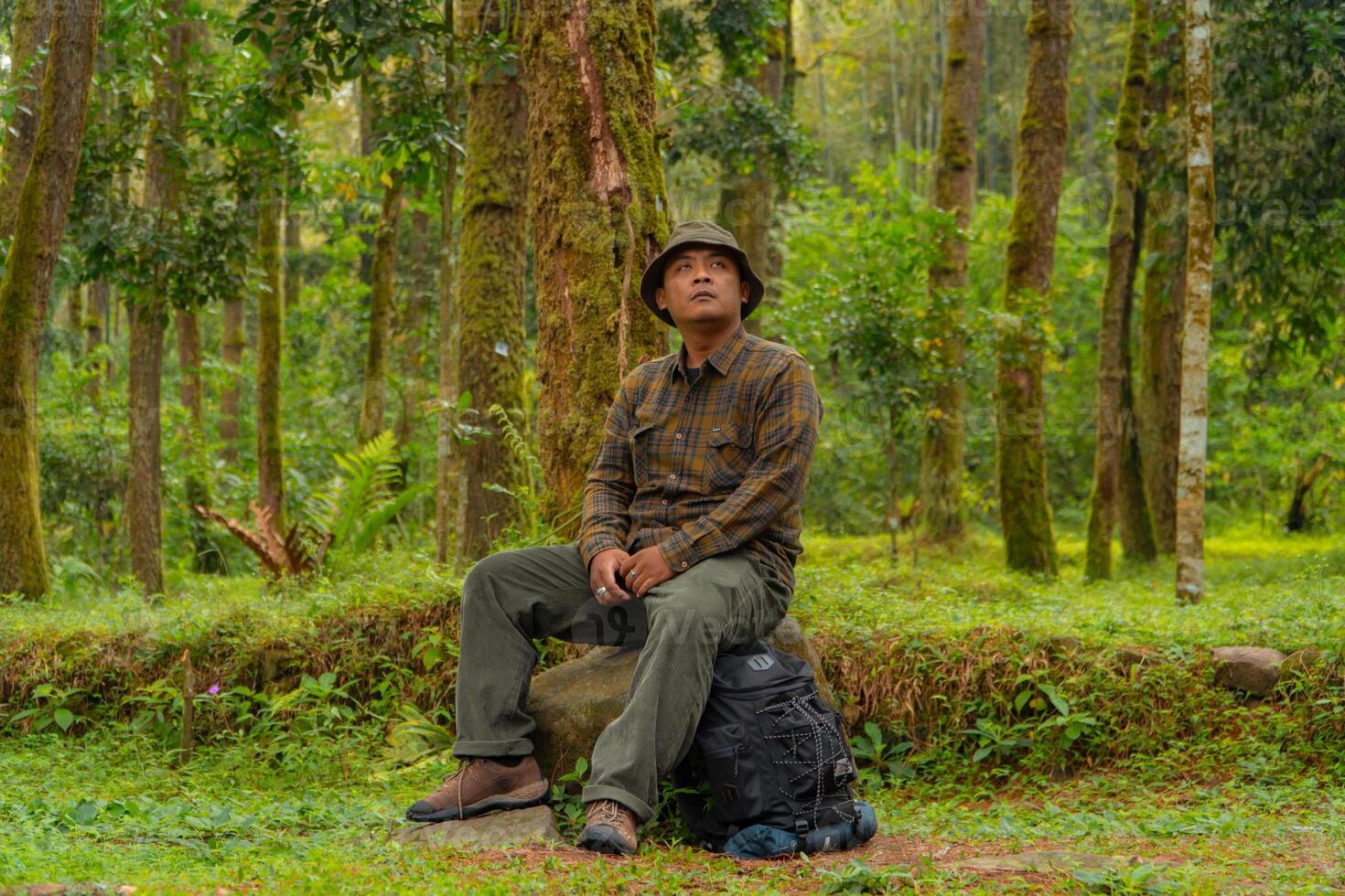 An adventurer is sitting on a large rock in a dense pine forest. An Asian man in a flannel shirt and bucket hat is sitting and resting while enjoying the tranquility of the beautiful nature. photo