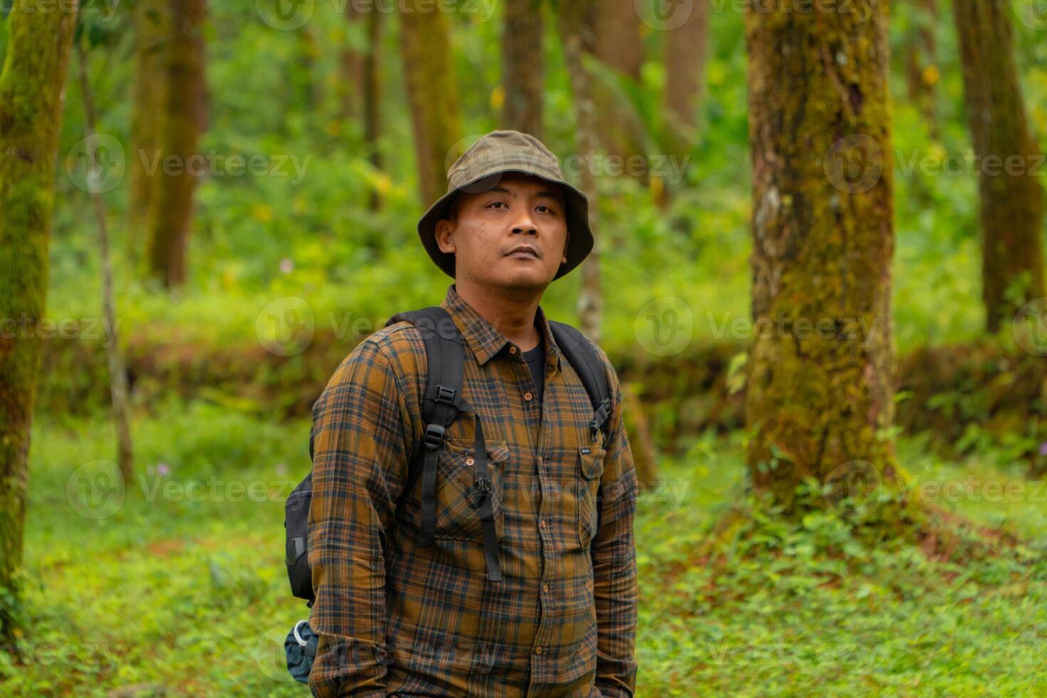 Half-body close-up of an adventurer in a dense and green pine forest. An Asian male traveler in a flannel shirt, bucket hat and backpack poses against a backdrop of dense pine trees. photo