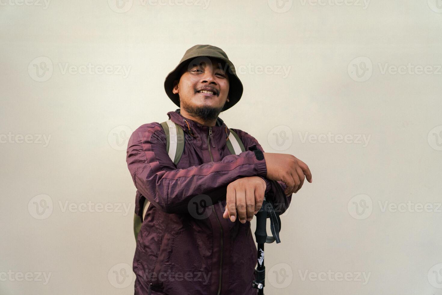 Half body portrait of adult Asian man with beard and mustache posing and wearing mountain climbing outfit with jacket, bucket hat, carrier bag and trekking poles isolated on beige background. photo
