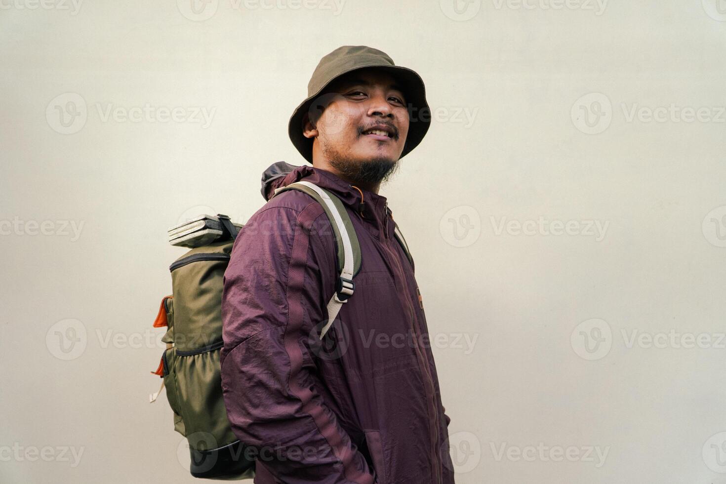 Half body portrait of adult Asian man with beard and mustache posing and wearing mountain climbing outfit with jacket, bucket hat, carrier bag and trekking poles isolated on beige background. photo