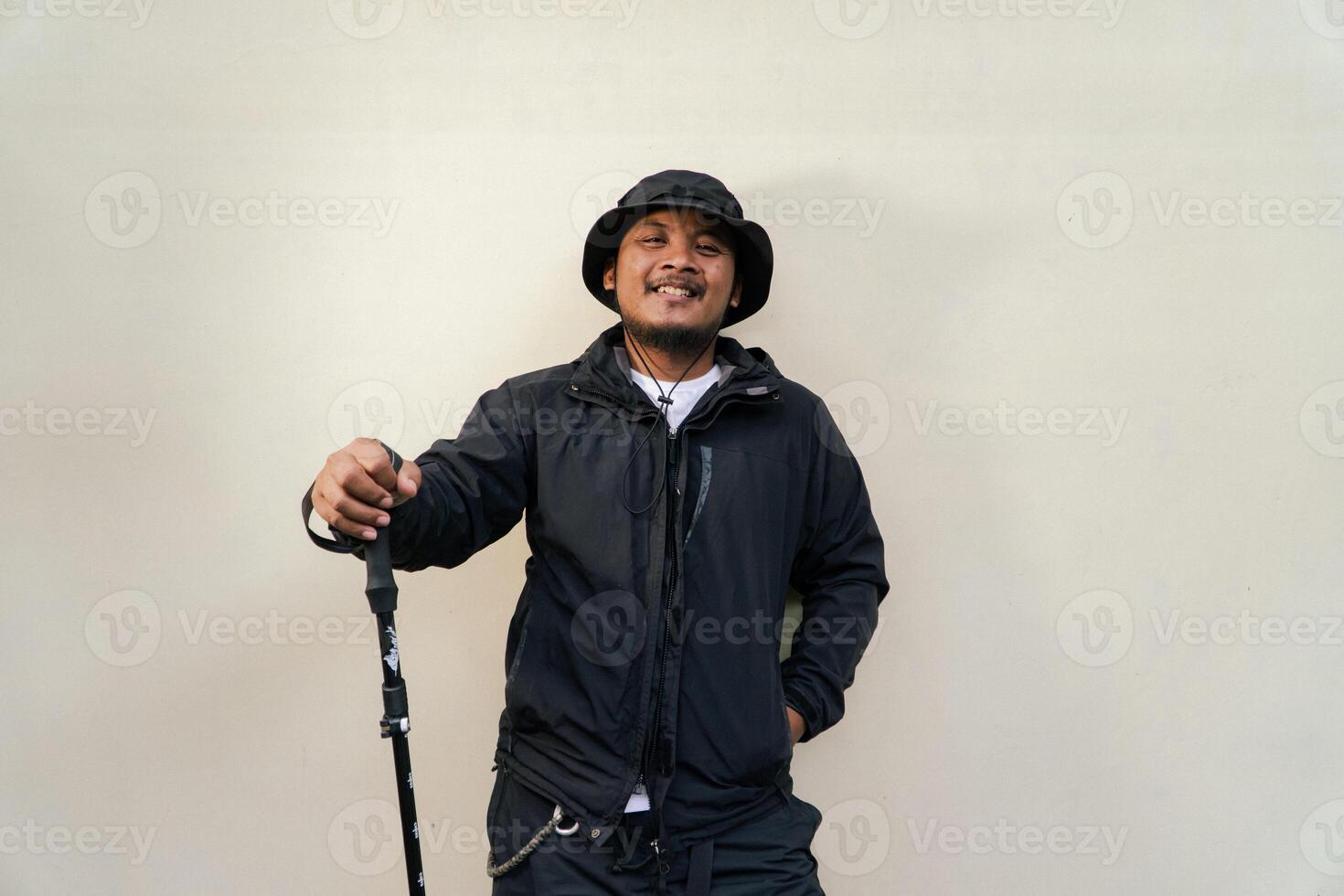 Photo of Asian man wearing adventuring outfit. Half body portrait of an adventurer with beard and mustache posing climber wearing jacket, boonie hat and trekking poles isolated on beige background