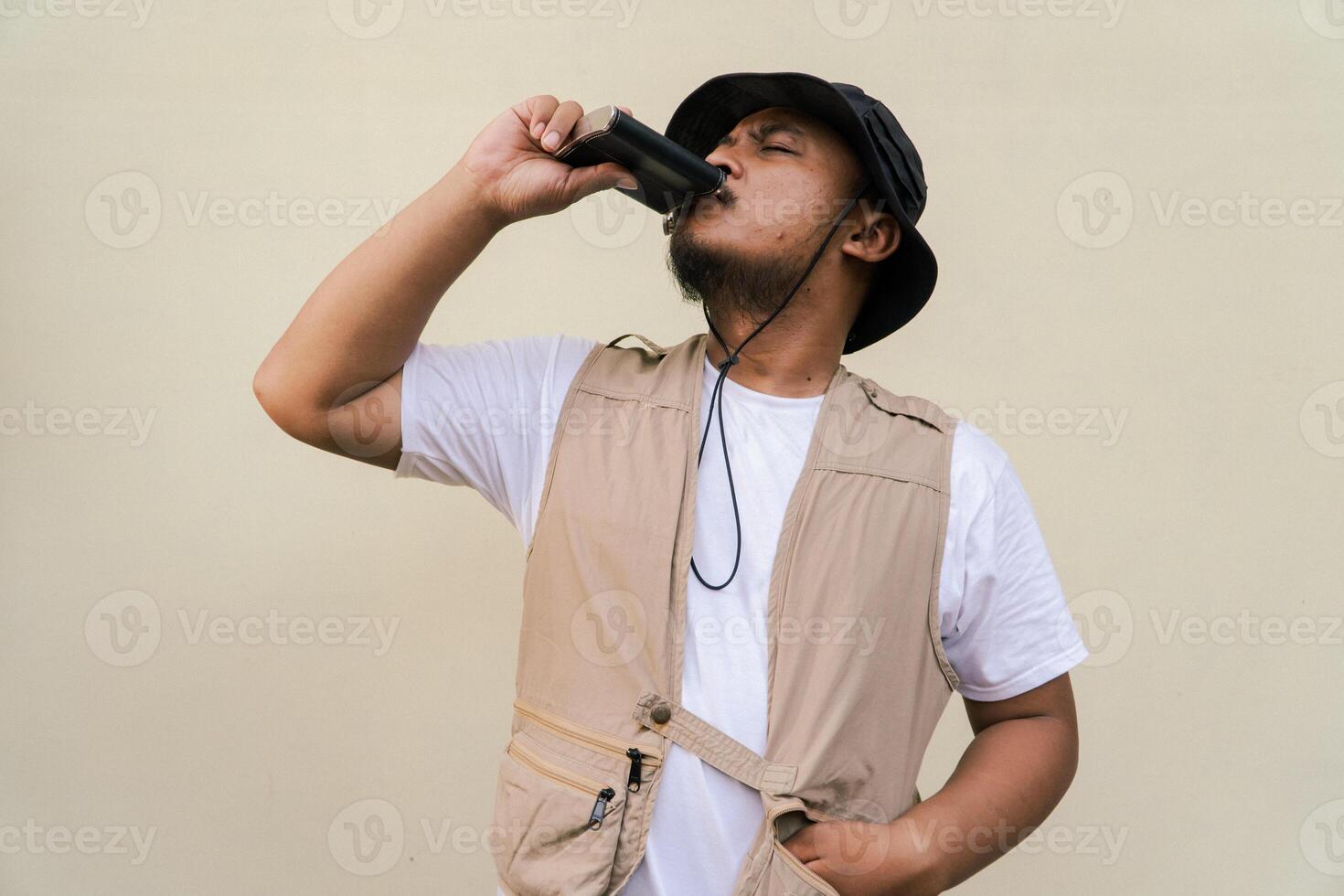Macho mature man with beard and mustache wearing safari outfit isolated on beige background. Half body portrait of an adult Southeast Asian man posing wearing a vest, bucket hat and hip flask bottle photo