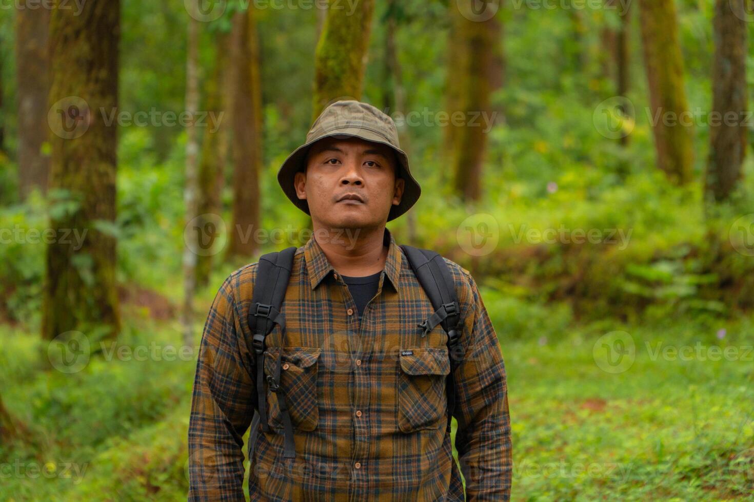 Half-body close-up of an adventurer in a dense and green pine forest. An Asian male traveler in a flannel shirt, bucket hat and backpack poses against a backdrop of dense pine trees. photo