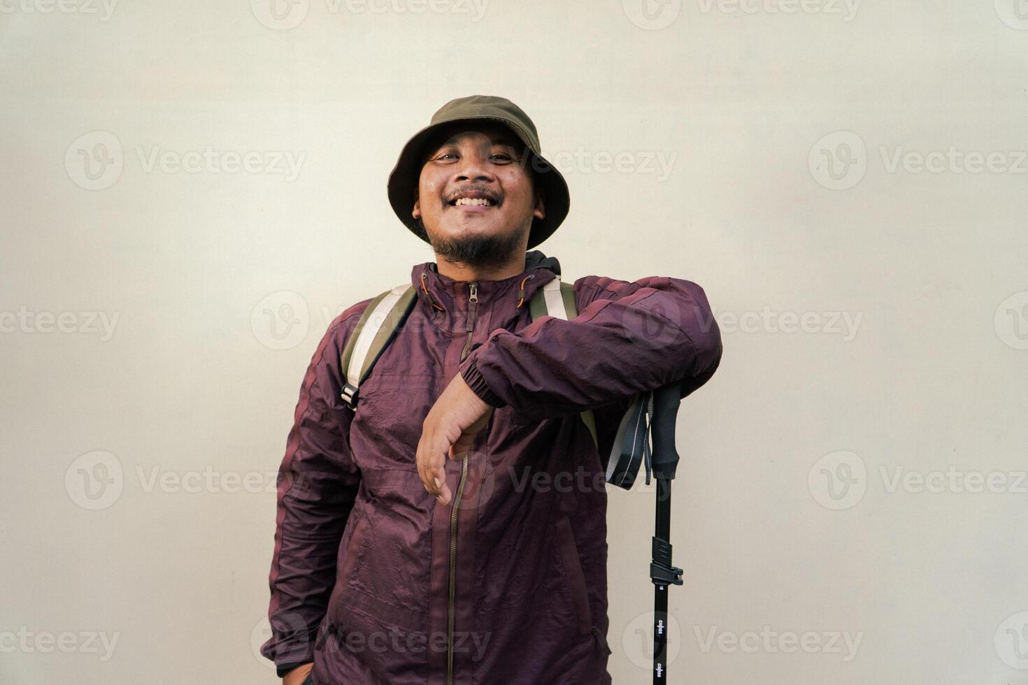 Adult Asian man wearing adventurer outfit and posing in various poses. An adventurer wearing maroon jacket, bucket hat, backpack and trekking pole isolated on beige background. photo