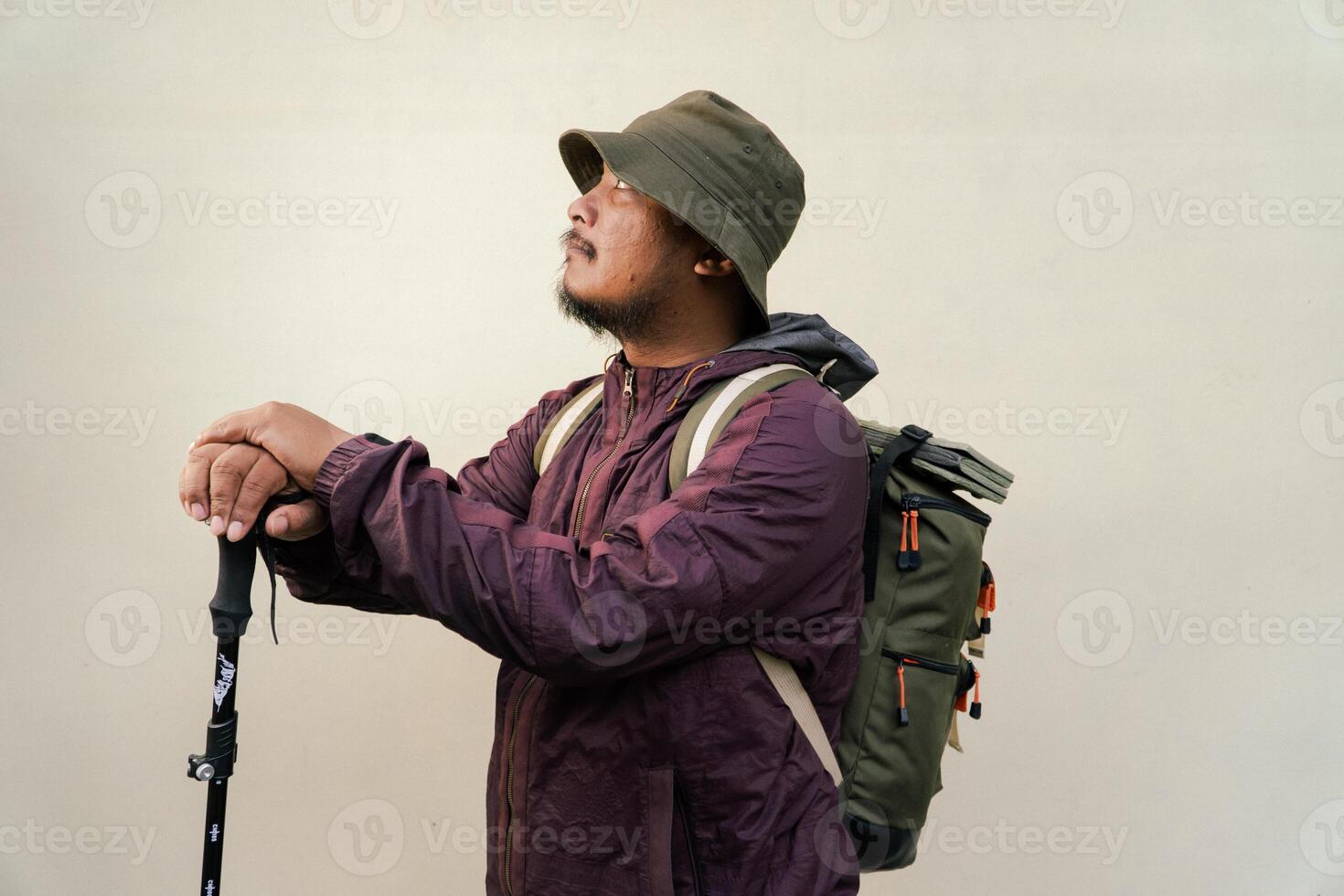 Adult Asian man wearing adventurer outfit and posing in various poses. An adventurer wearing maroon jacket, bucket hat, backpack and trekking pole isolated on beige background. photo