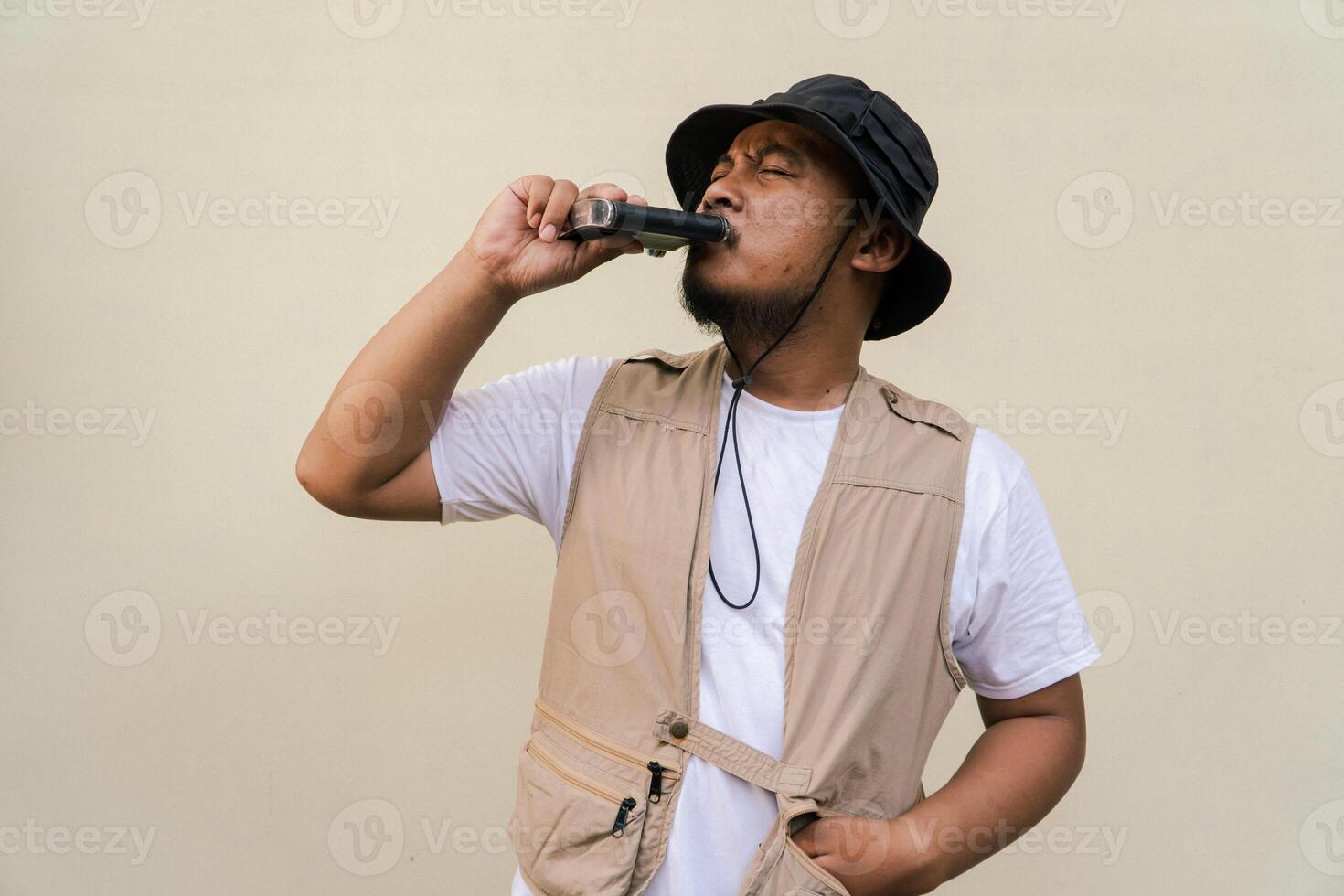 Macho mature man with beard and mustache wearing safari outfit isolated on beige background. Half body portrait of an adult Southeast Asian man posing wearing a vest, bucket hat and hip flask bottle photo
