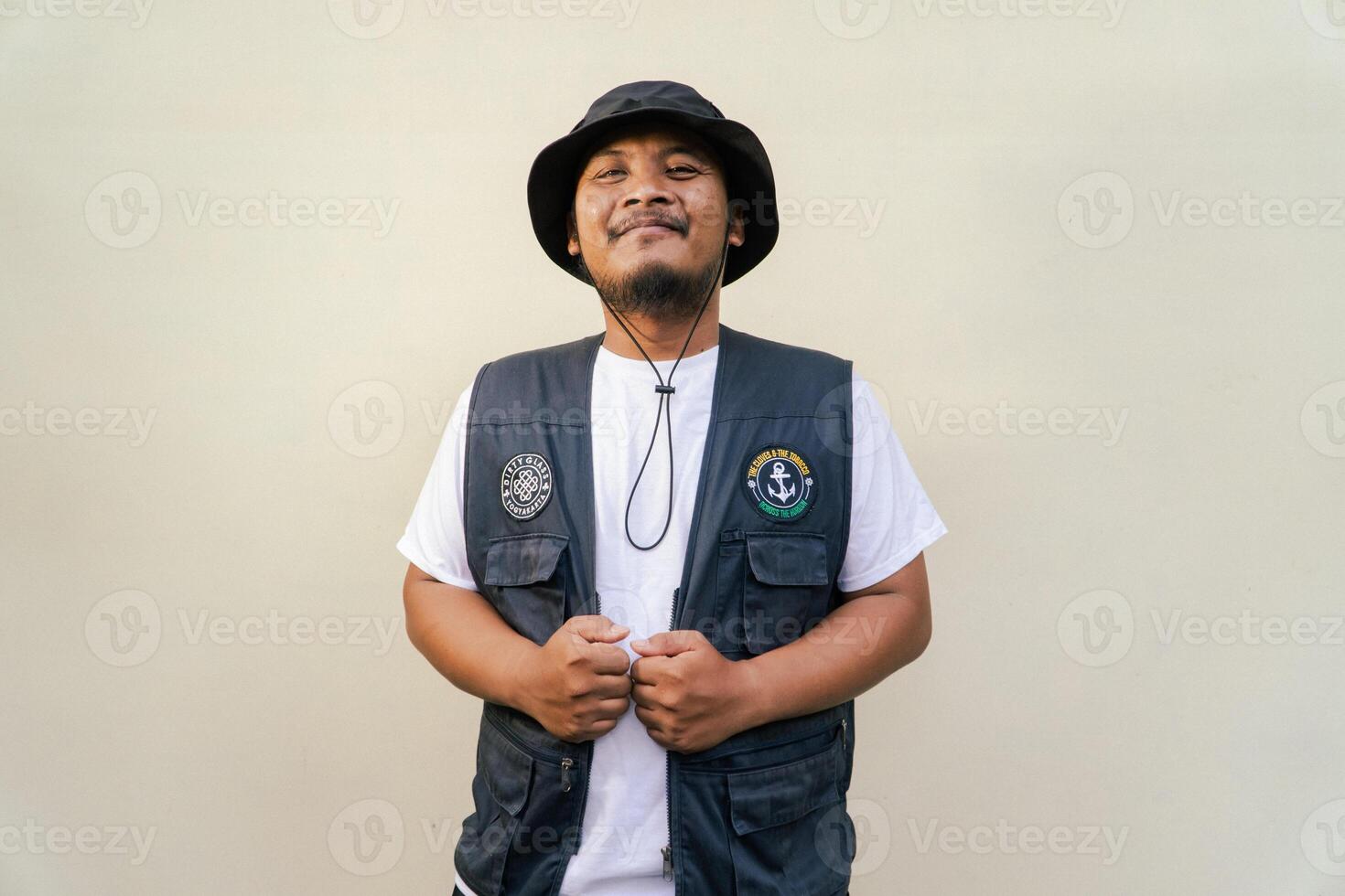 Asian adult man wearing safari suit isolated on beige background. Half body portrait of adult Southeast Asian man posing wearing vest, bucket hat and white t-shirt photo