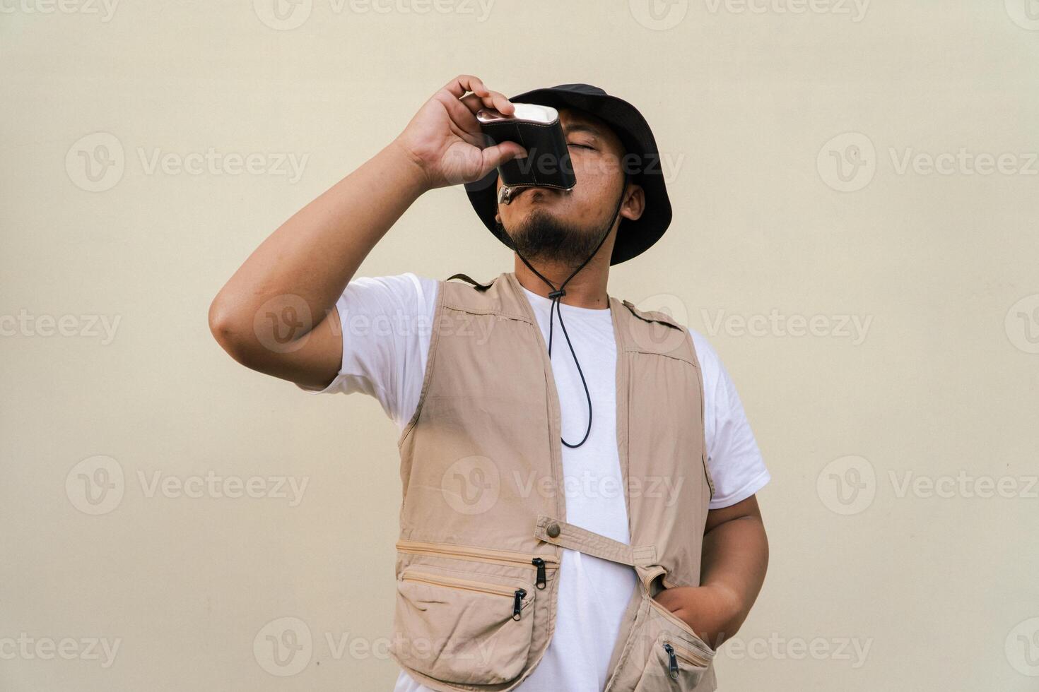 Macho mature man with beard and mustache wearing safari outfit isolated on beige background. Half body portrait of an adult Southeast Asian man posing wearing a vest, bucket hat and hip flask bottle photo