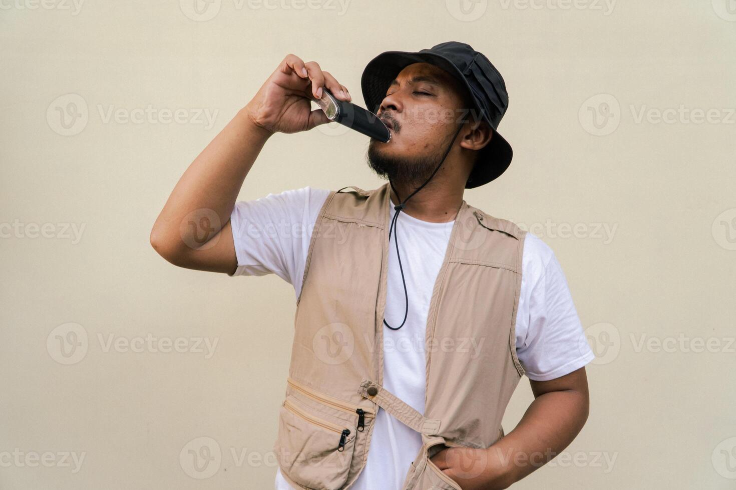 Macho mature man with beard and mustache wearing safari outfit isolated on beige background. Half body portrait of an adult Southeast Asian man posing wearing a vest, bucket hat and hip flask bottle photo