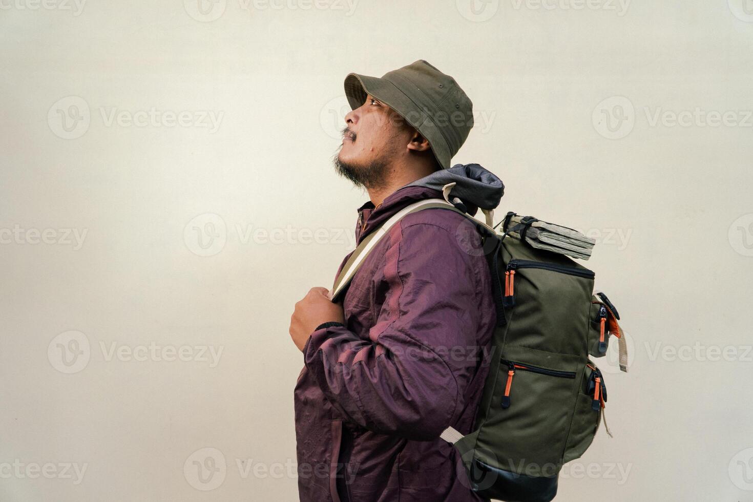 Half body portrait of adult Asian man with beard and mustache posing and wearing mountain climbing outfit with jacket, bucket hat, carrier bag and trekking poles isolated on beige background. photo