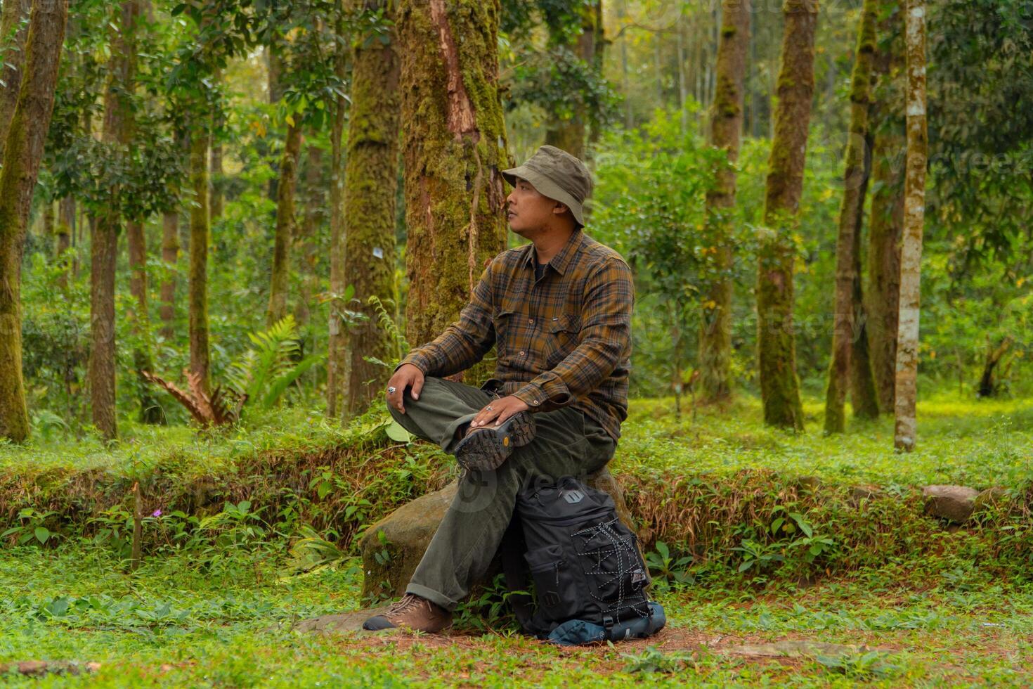 An adventurer is sitting on a large rock in a dense pine forest. An Asian man in a flannel shirt and bucket hat is sitting and resting while enjoying the tranquility of the beautiful nature. photo